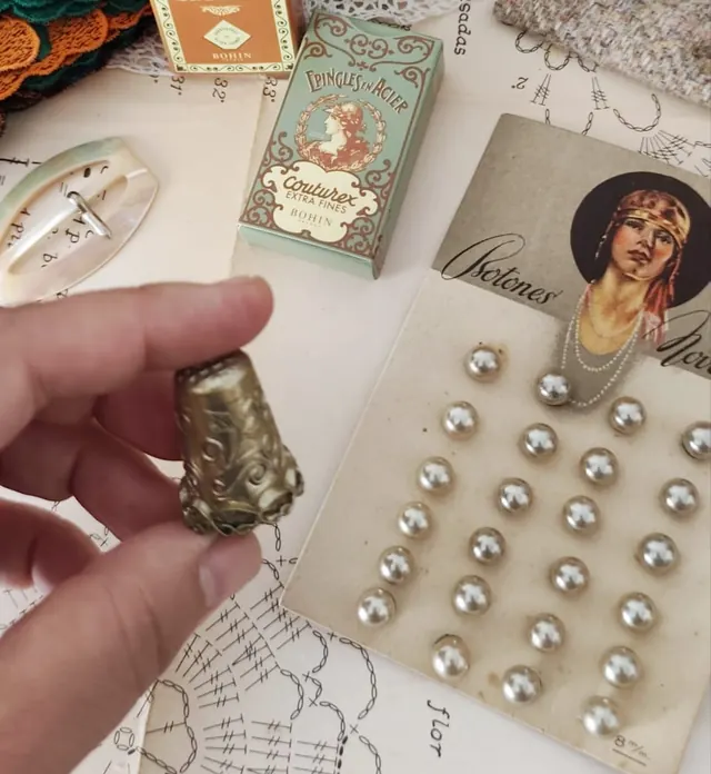 Dedal antiguo en la mano junto a alfileres Couturex de Bohin y botones de perla en cartón